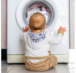 Baby sitting on floor in front of a washing machine full of clothing.